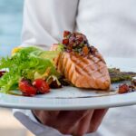 waiter holds a plate of grilled smoked salmon with lettuce, tomato, pepper