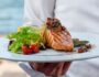 waiter holds a plate of grilled smoked salmon with lettuce, tomato, pepper